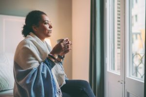 Woman wearing blanket and drinking hot tea looks out of the window