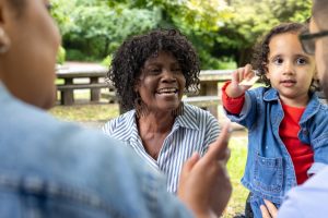 Close-up over-the-shoulder shot of a grandmother and toddler with two parents enjoying a picnic. The main focus is on the smiling grandmother, the toddler beside her reaching towards her father with a piece of food in hand.