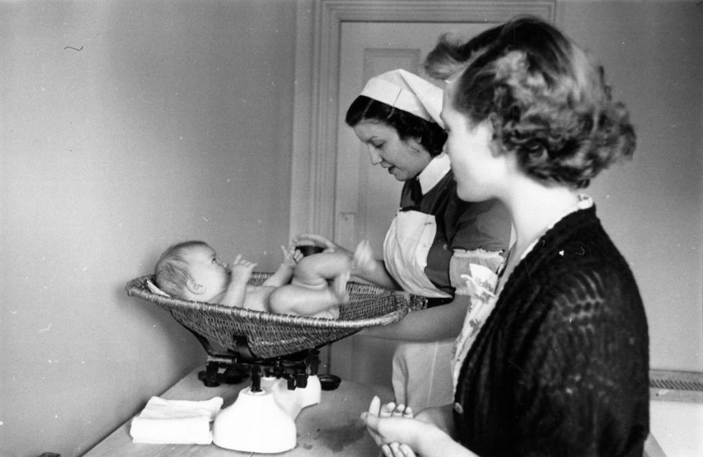 Black and white photo of nurse weighing a baby, 1950s.