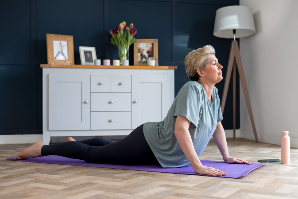 A full shot of a woman who is living with Breast Cancer doing a cobra position on a purple yoga mat. She is wearing casual gym attire in her home in the North East of England. Videos are available similar to this scenario.